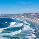 A breathtaking aerial shot of San Diego's coastline, capturing waves and cityscape under a clear sky. Blog post about taking a trip to San Diego